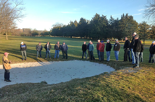 17 people on a golf course listening to a speaker standing in a sand trap.